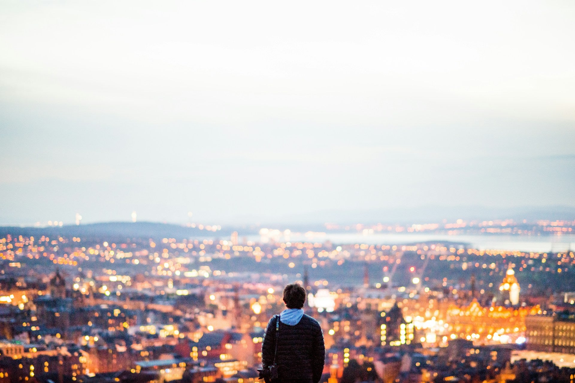 Our story banner: a person stands on a high viewpoint, looking out over a city at dusk with thousands of warm, blurred lights and a wide pale sky, creating a calm, reflective mood.